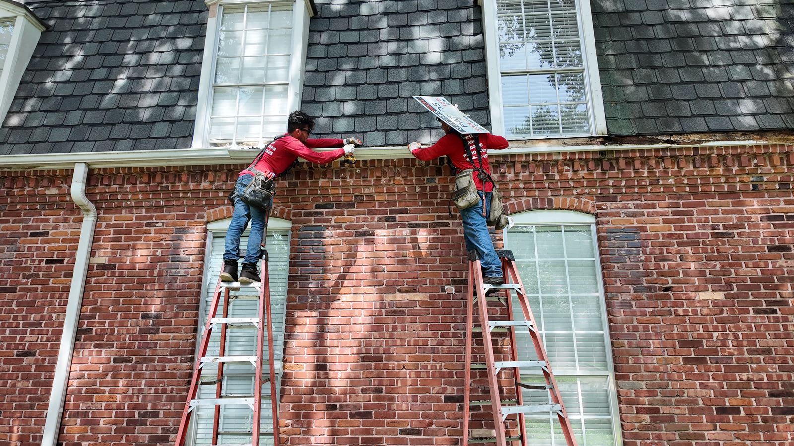 GuttersTech technicians carrying a fascia board up a ladder while servicing the roofline of a brick home